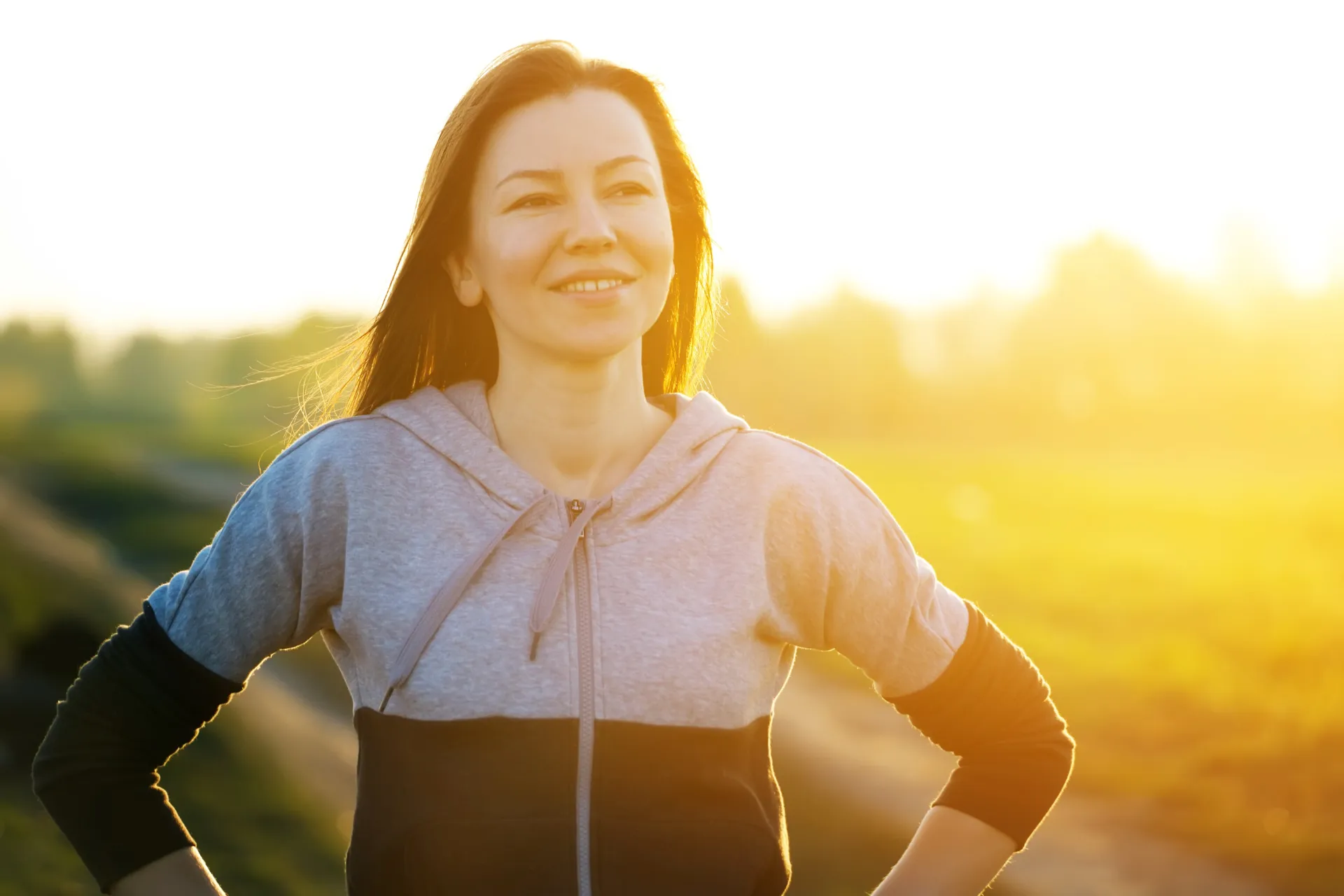 Smiling woman in a hoodie standing outdoors at sunset after a workout